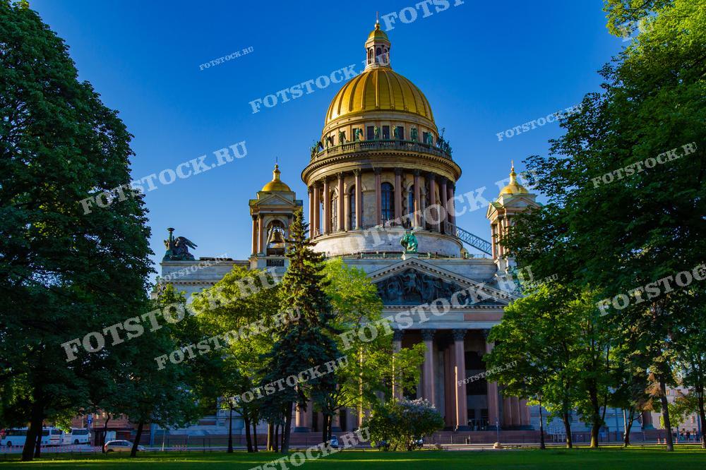 На фотографии запечатлен Исаакиевский собор в Санкт-Петербурге, окружённый зелёными деревьями под ярким солнечным светом. Золотистый купол сияет на фоне синего неба, создавая величественный и торжественный образ. Это изображение  подходит для туристических буклетов, образовательных материалов, и как украшение для интерьера любителей архитектуры.